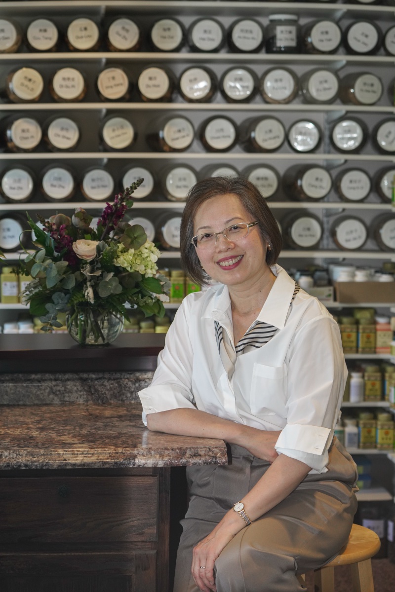 Dr. Hedy Chen seated at the clinic's herbal pharmacy, shelves of labeled Chinese herbs behind her